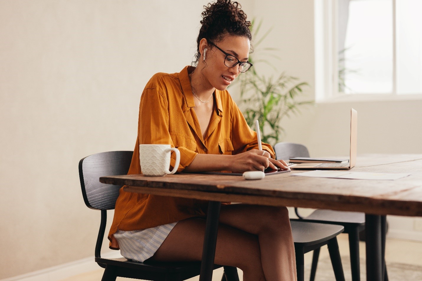 A person sitting at a wooden table with a cup of coffee, their airpods in, and a laptop off to the side. They are writing on a tablet with their stylus.