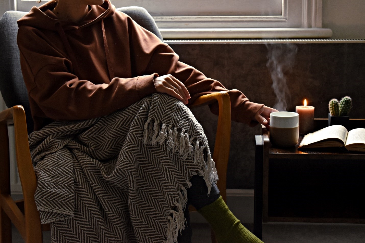 A person reaching for a steaming mug next to a book. The person is wearing a large, brown hoodie with a blanket over their lap.
