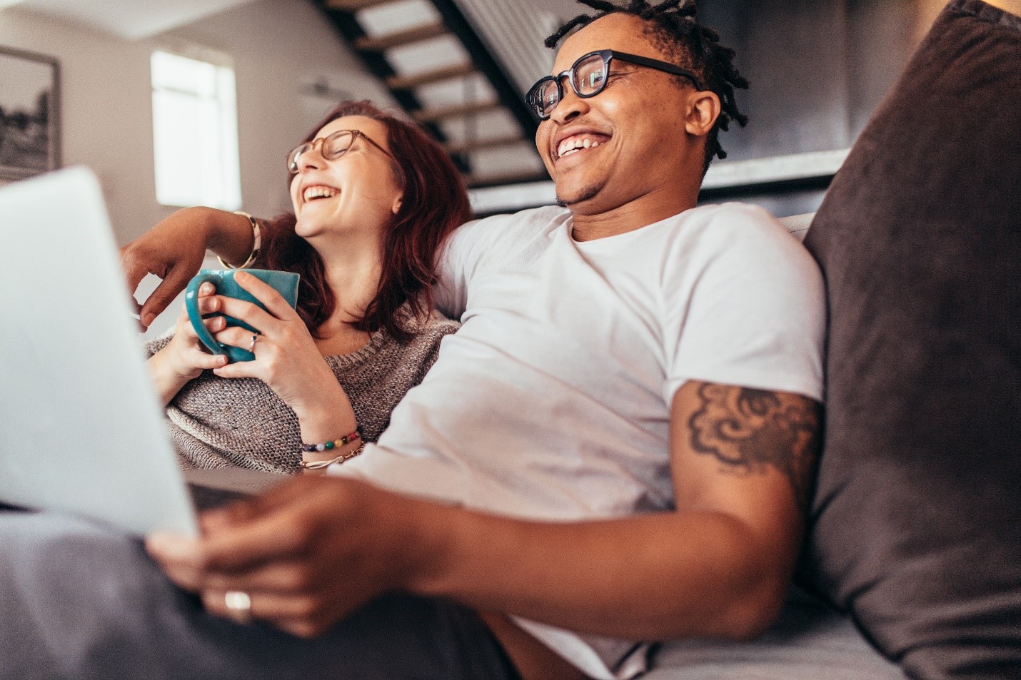 Two people sitting comfortably. They are laughing and in front of a laptop.