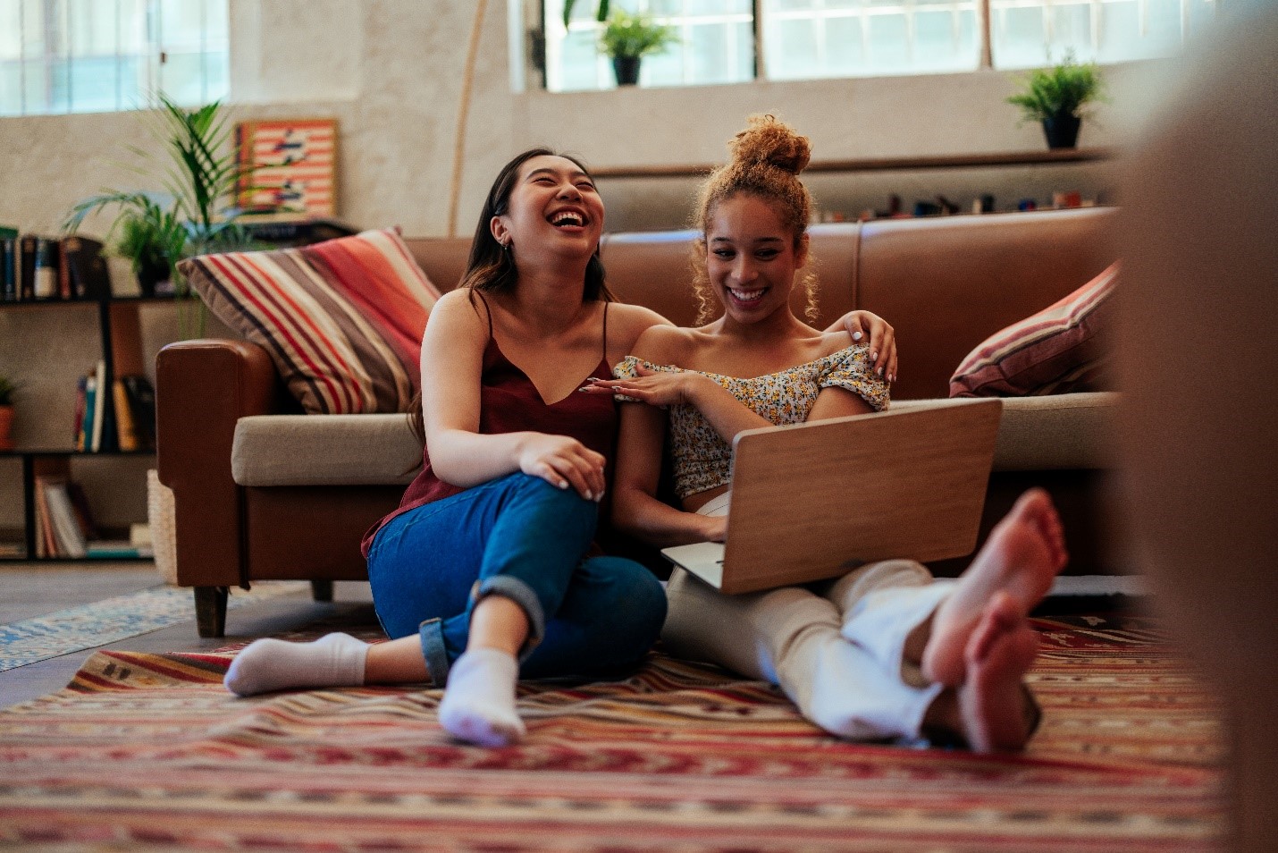 Two femme people sitting on the floor in front of a couch and laughing while looking at a laptop.