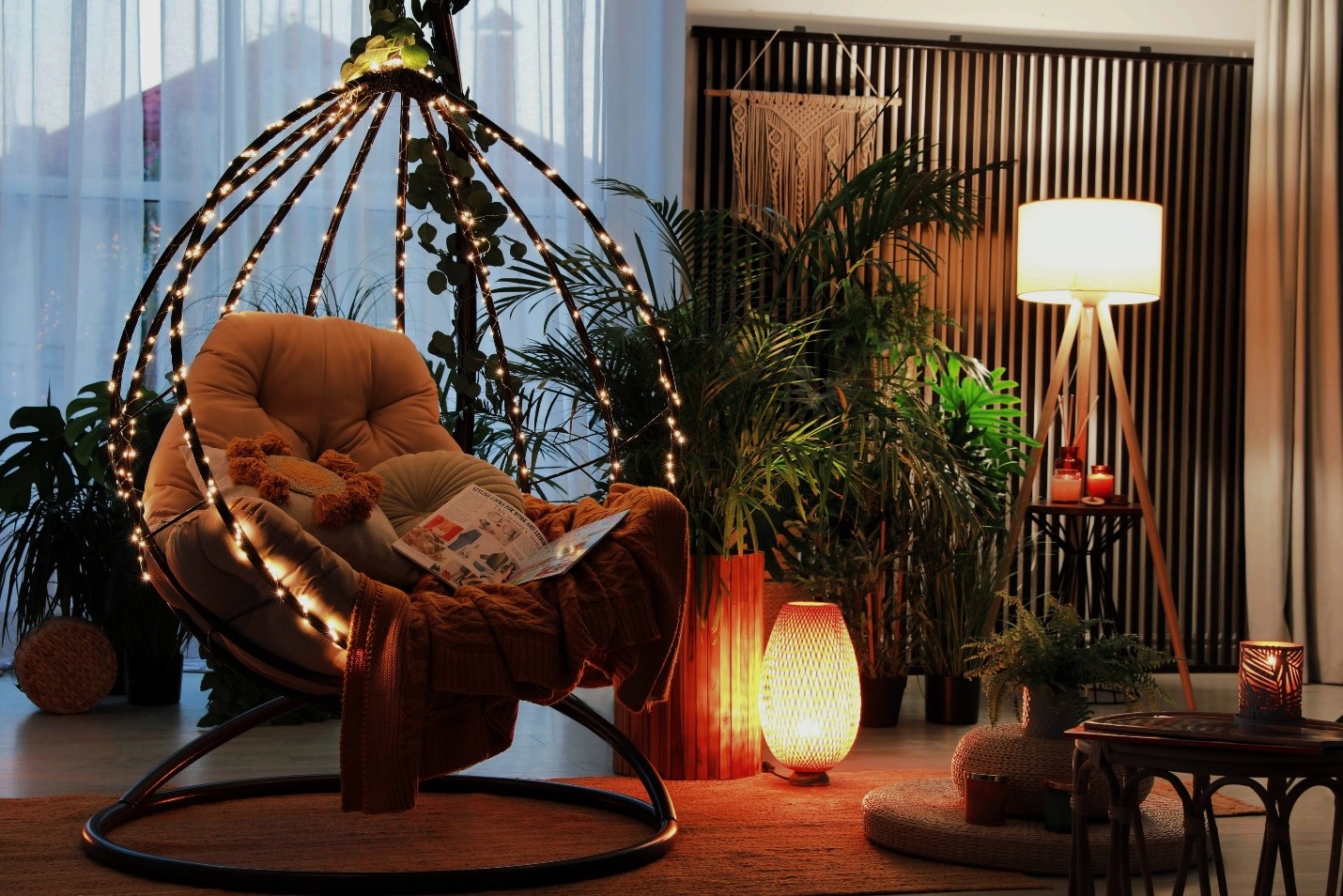 An interior space shot of a hanging chair decorated with soft fairy lights. It's covered in blankets and soft pillows, a magazine is left open on top. There is a soft orange hued light on the floor and a larger light behind that. On a table to the right, there is a lit candle.