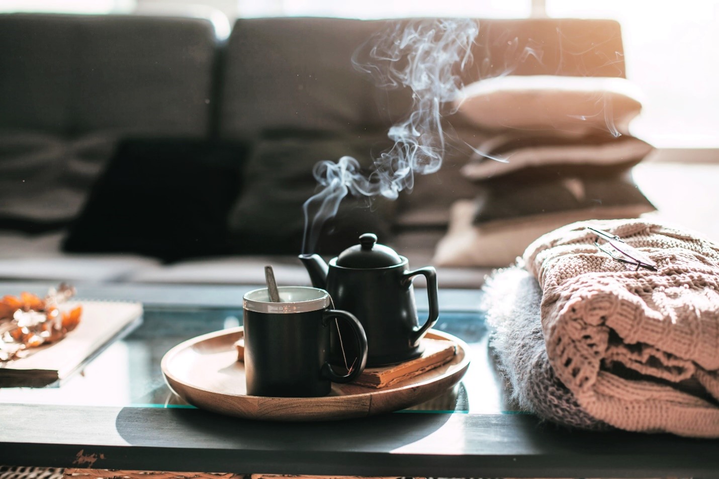 A black teapot is steaming as it sits next to a black tea cup. Both are positioned on a wooden plate on top of a glass coffee table. There are blankets stacked on the side.