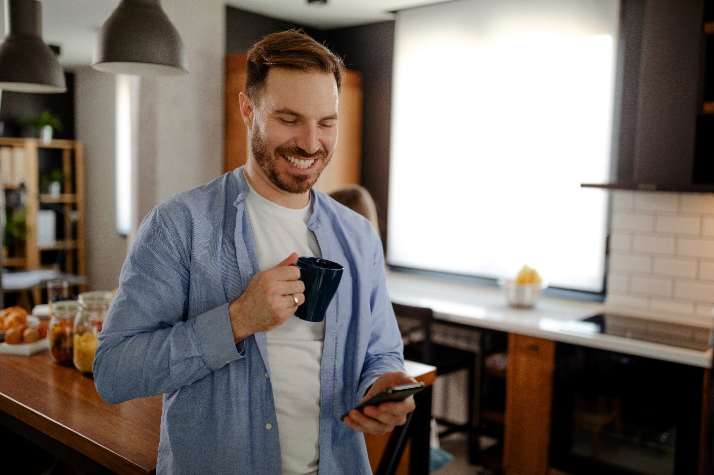 A man standing in his kitchen with a coffee cup in one hand and his phone in the other. He's smiling down at his phone.