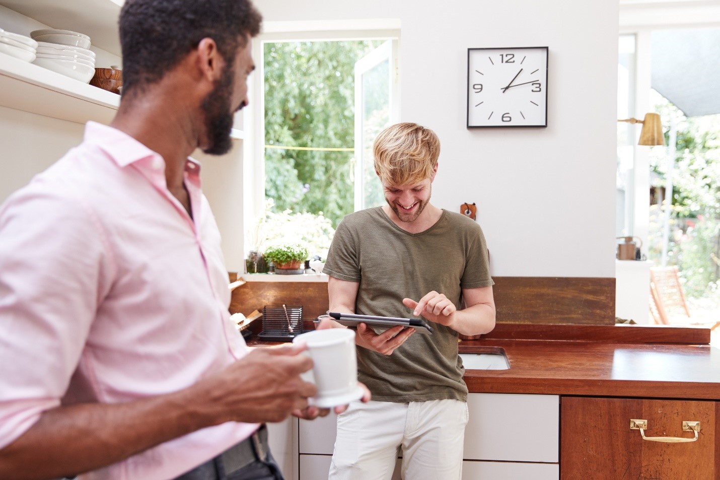 Two male presenting people are standing in a kitchen together and smiling. One has coffee in their hand. The other is tapping on a tablet.