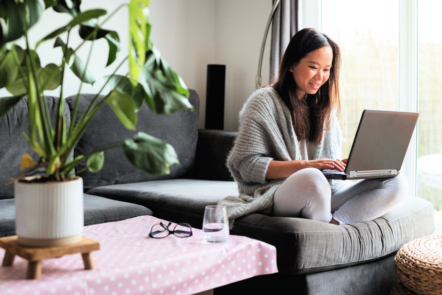 A femme person working on her laptop on the edge of her couch. There is a glass of water and glasses next to her. 