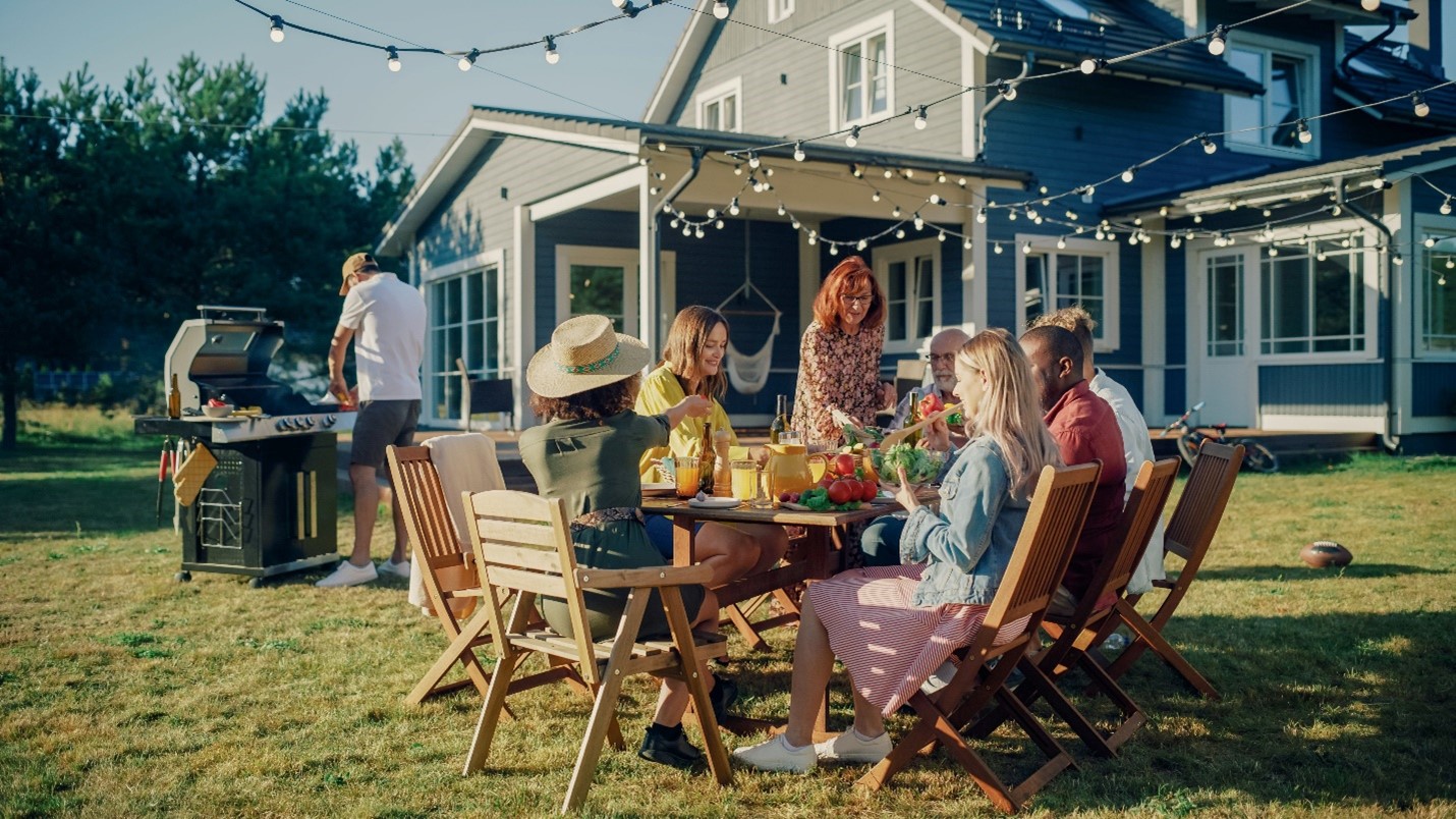 An outdoor party in a large backyard, decorated with hanging string lights. On the left, a man stands by a grill, his face hidden behind a hat. A woman is standing to serve a salad to guests.