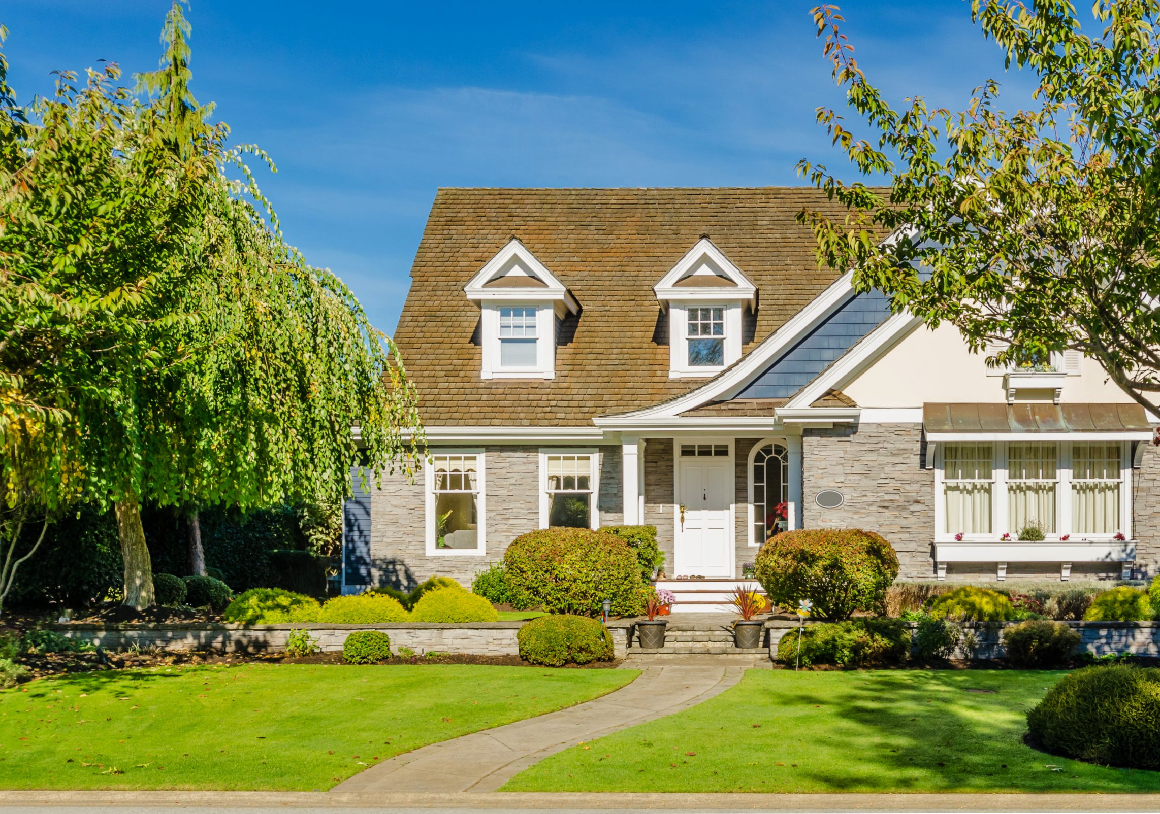 A home with large trees, landscaping, rock retaining walls, and a bright exterior on a sunny day.