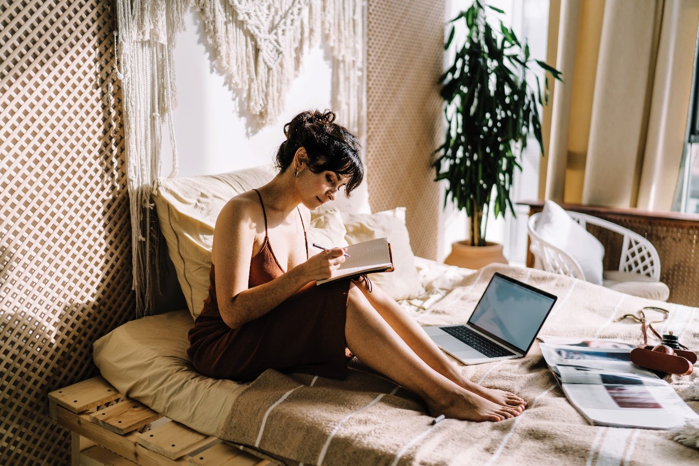 A woman writing in her notebook as she sits barefoot on her bed.