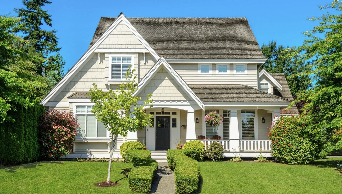 A large beige home with a covered porch, surrounded by trees and shrubs.