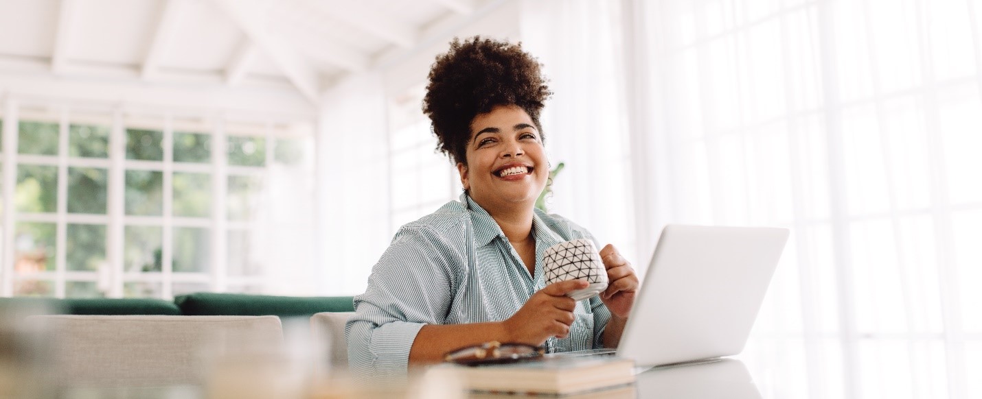 A Black woman holds a coffee cup in front of her laptop. The room is filled with light and she's smiling at someone just off camera.