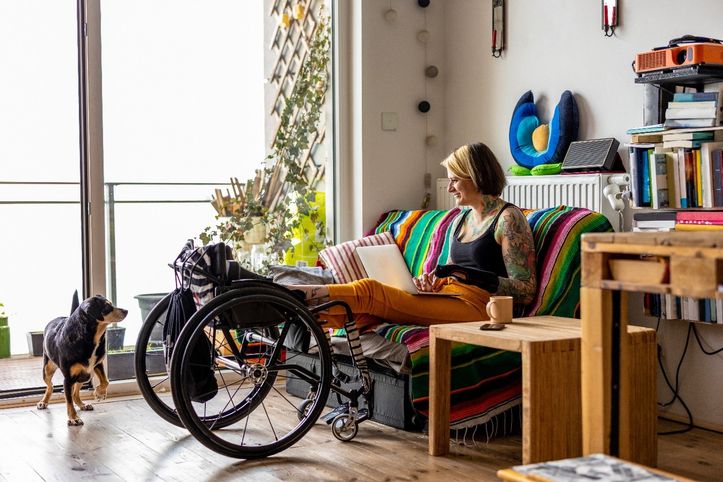 A woman sits on her couch with her laptop on her lap. Her legs are up on her wheelchair as she smiles at her dog.