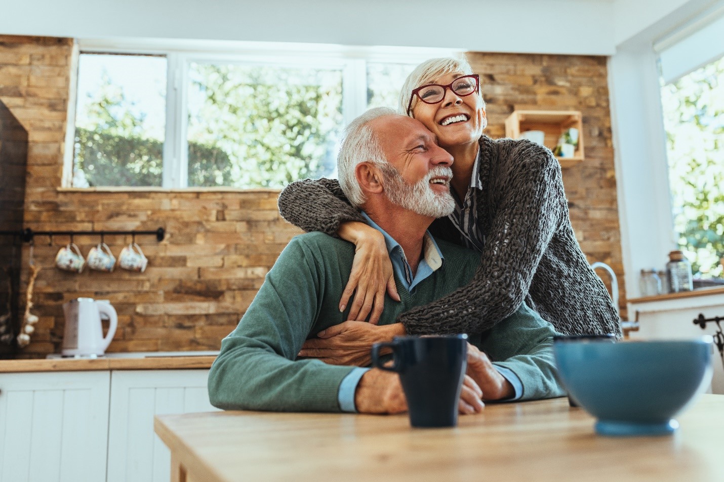 An older couple hug in their kitchen.