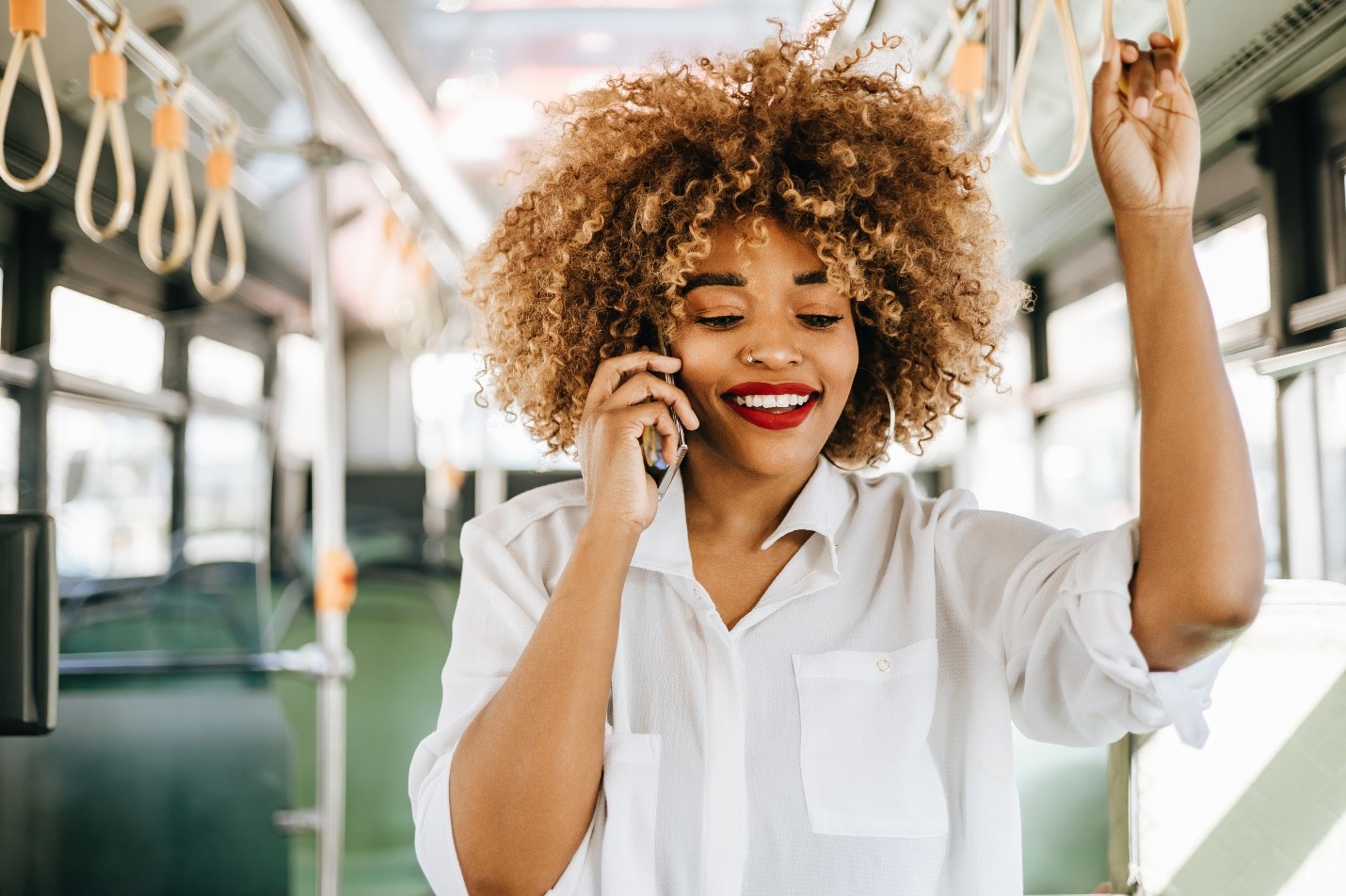 A young femme person with a curly afro and red lipstick holds onto the train as she speaks on her cell phone.