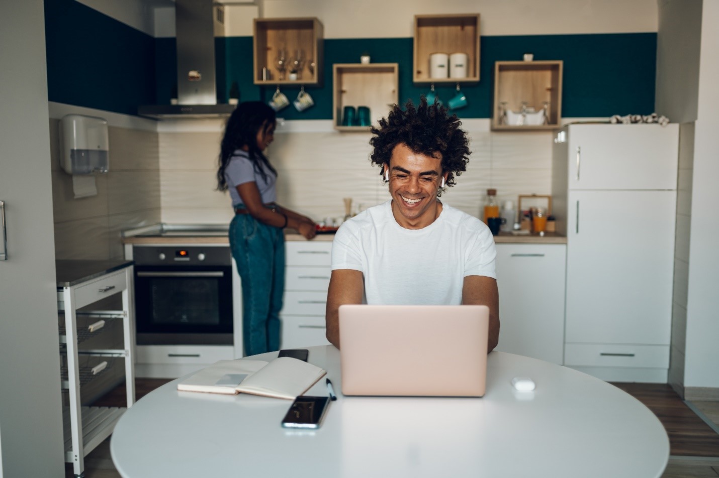 A man on his laptop in the kitchen while his partner cooks in the background.