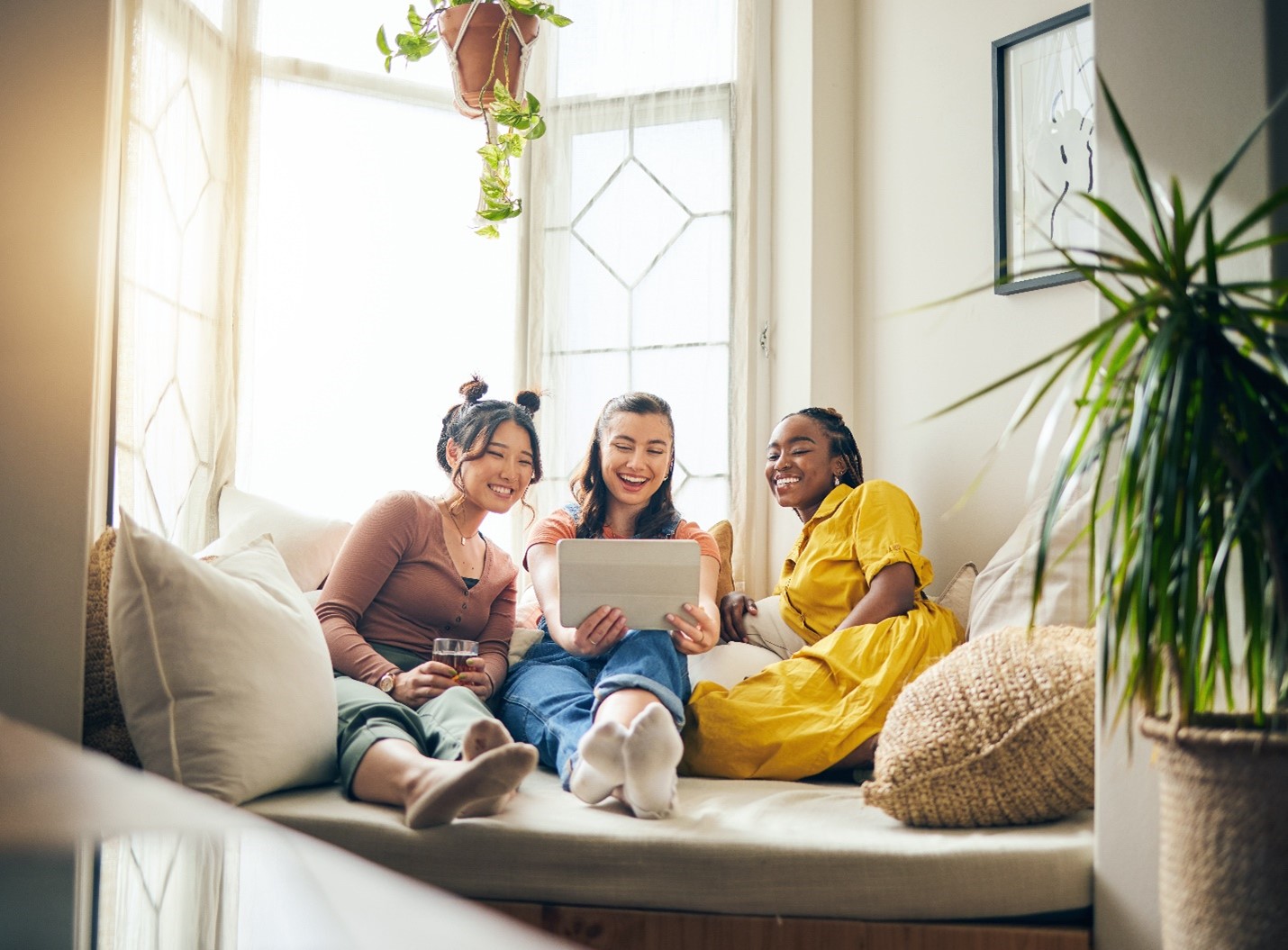 Three young people of different nationalities smiling at a tablet together. They're sitting on a couch together in their home.