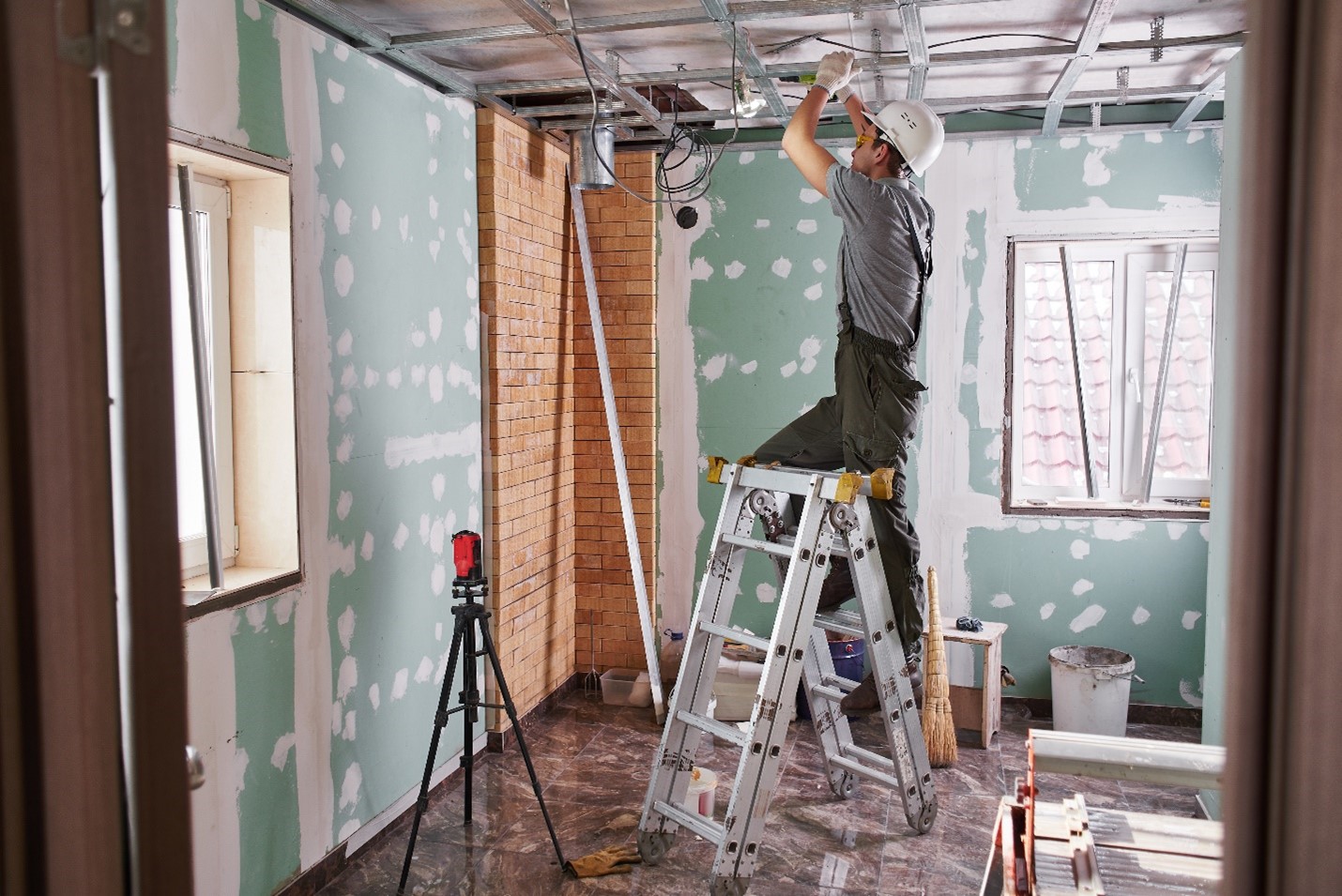 A man stands on a small step ladder, drilling in framing for a room's ceiling.