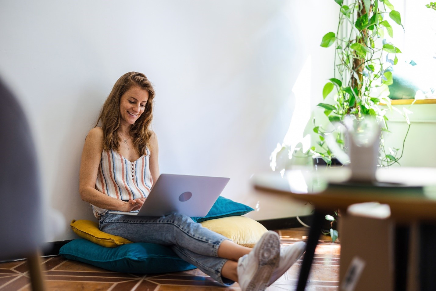 A smiling woman sits on pillows on the floor with her laptop open in front of her.