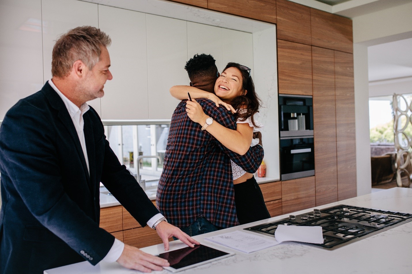 Multiracial couple hugging in front of their real estate professional inside of their new home.
