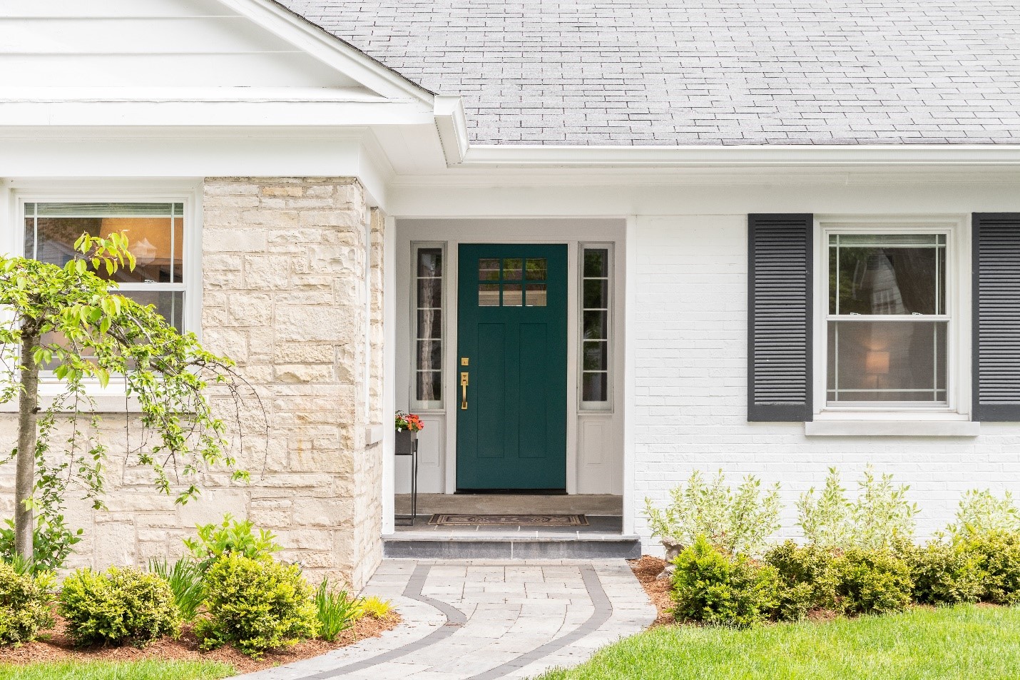 The front walk to a white brick home. The front door is teal with gold accents. A light is on and visible in the front window.