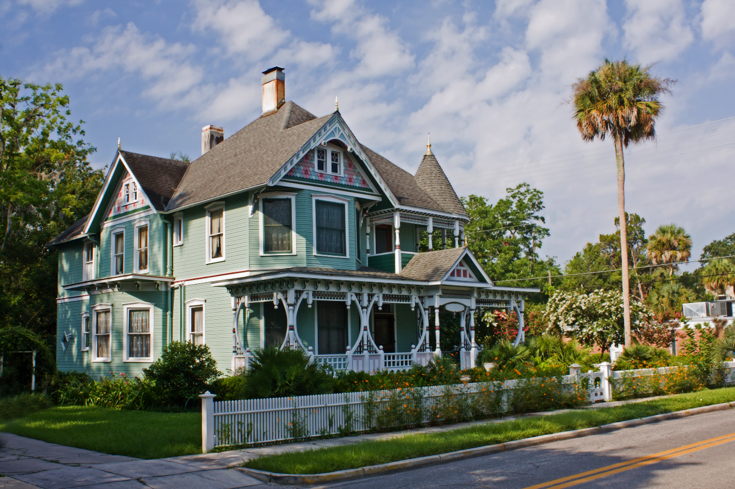 A large turquoise Victorian home with a wrap around porch with decorative pillars and intricate gables.