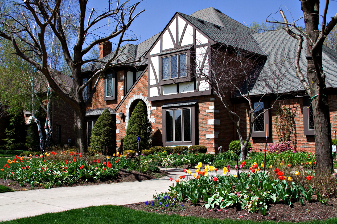 A large Tudor home with an arched stone front door, brick walls, and paneling detailing surrounding the dormer's windows.